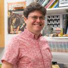A smiling man wearing glasses and a red patterned shirt stands in a colorful office or lab space. Behind him are shelves filled with books, framed artwork, and quirky items.